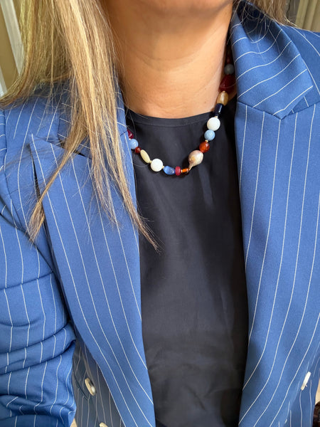 A close-up of a person wearing a colorful beaded necklace with red, blue, white, and beige beads, styled over a black top and a blue pinstripe blazer. Inamullumani 