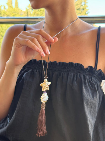 A person wearing a black off-the-shoulder top holds up a bohemian-style necklace featuring a gold-toned starfish, a white shell bead, and a brown tassel.