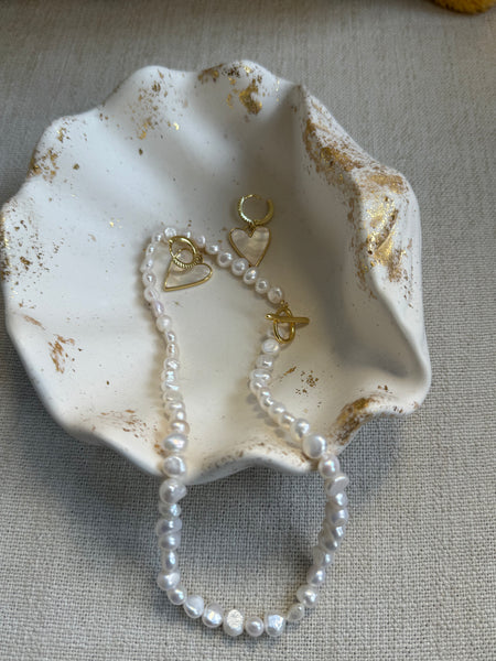 White Pearl  necklace and matching with gold heart-shaped earrings, displayed in a distressed white shell dish on a textured beige fabric background.